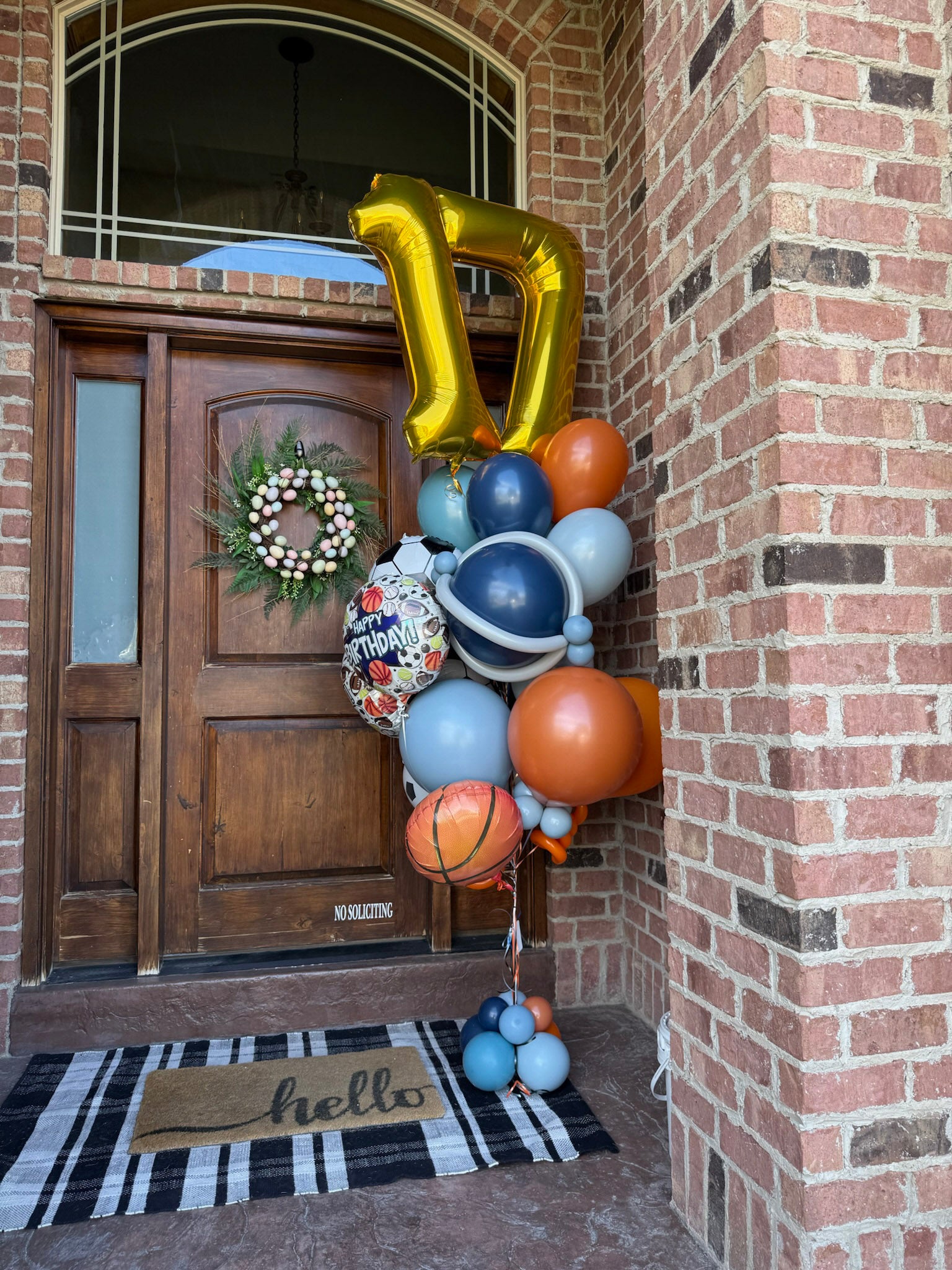 Decorative, custom helium balloon arrangements sits on the front steps of a home in Utah County after being delivered. It features a gold 17 jumbo numbers with orange, blue, and sports balloons.