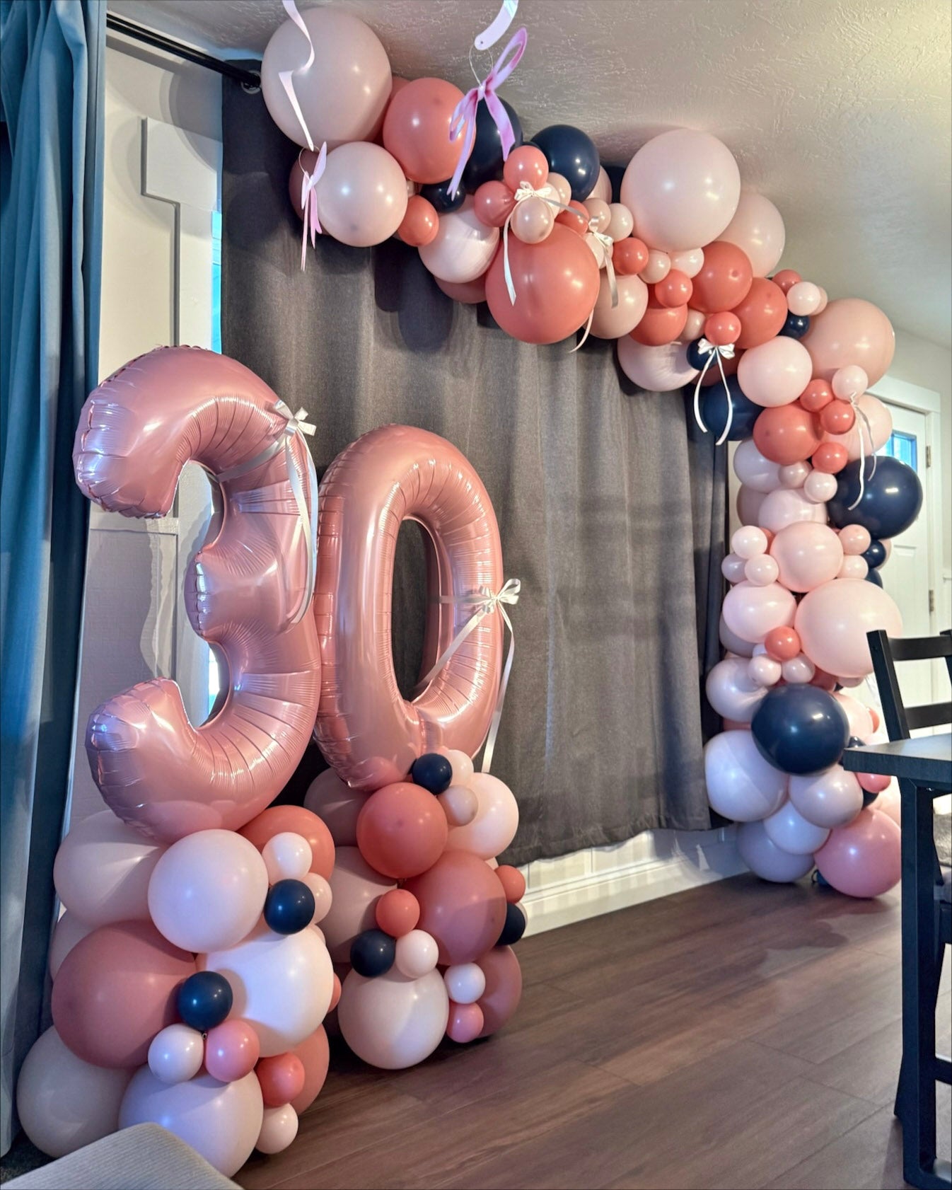 Balloon garland arches up and over a window in a living room featuring light pink, rose pink, and navy balloons. Additional balloon columns on the side of the room show a pink 30 for a 30th birthday. Additional ribbons can be seen on and throughout the balloons.