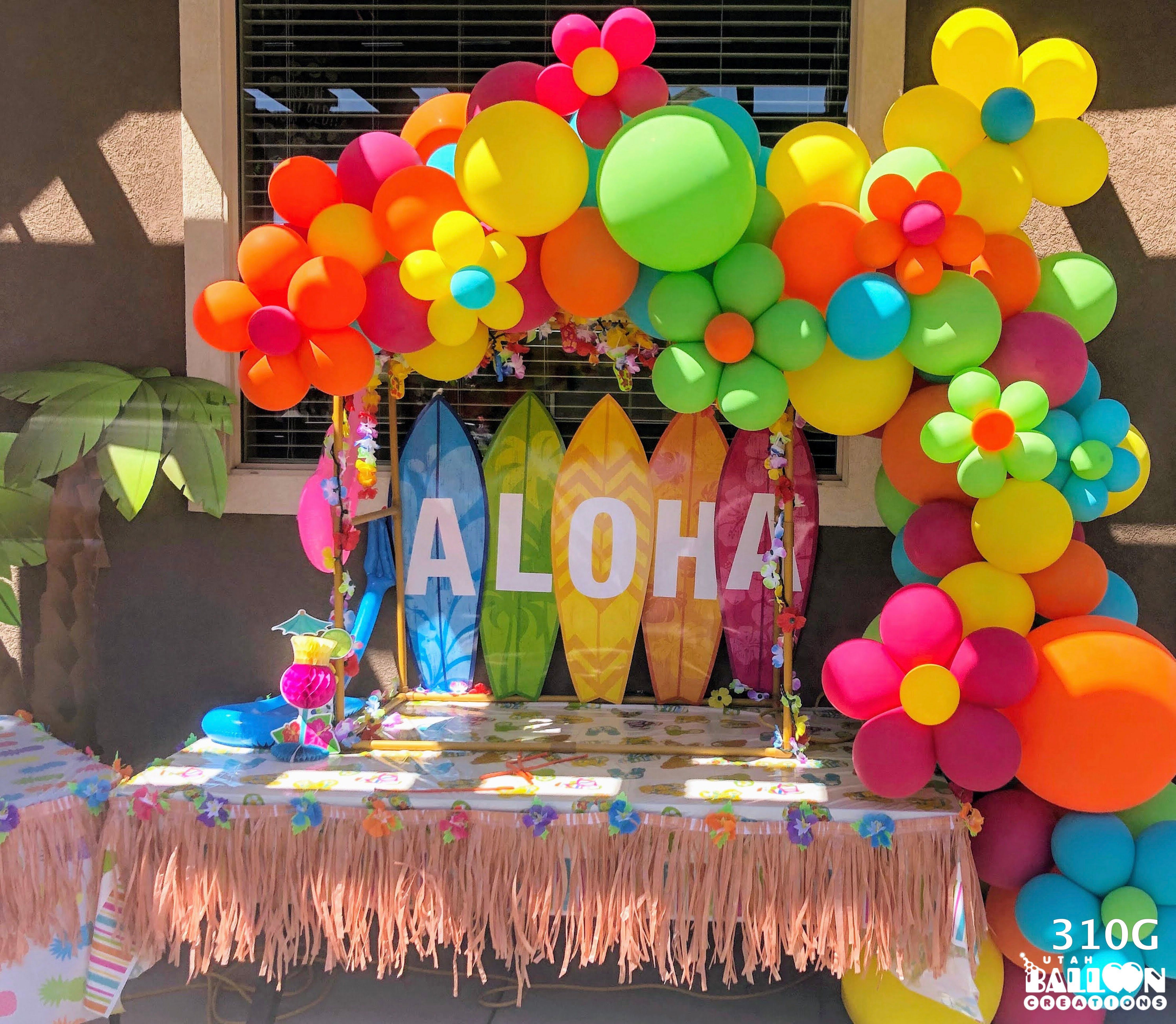 Decorative setup with colorful balloons, flowers, and 'ALOHA' sign on a table.
