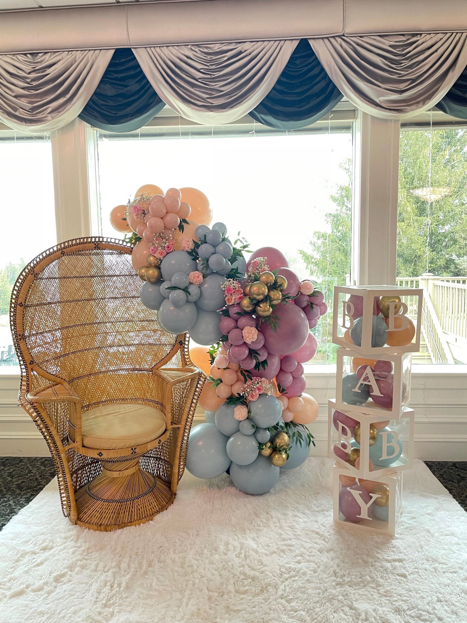A light blue, purple, and blush balloon garland winds up the side of a peacock chair at a baby shower. Baby blocks filled with balloons are on the side.