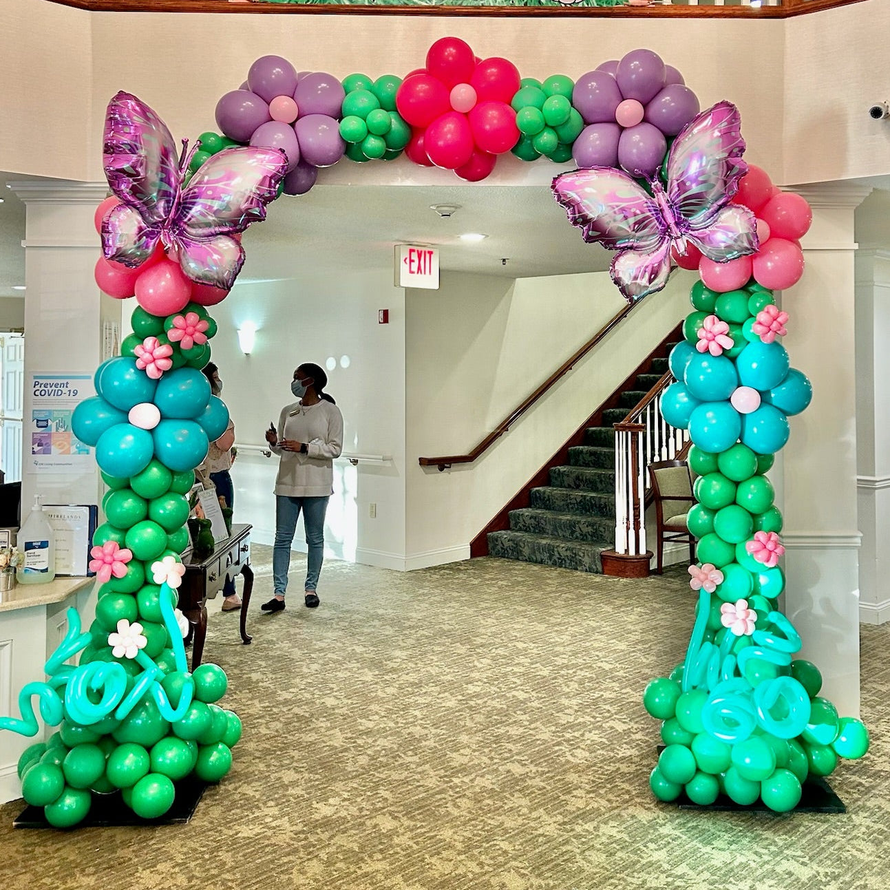 Decorative balloon arch with colorful flowers and butterflies in a building lobby.