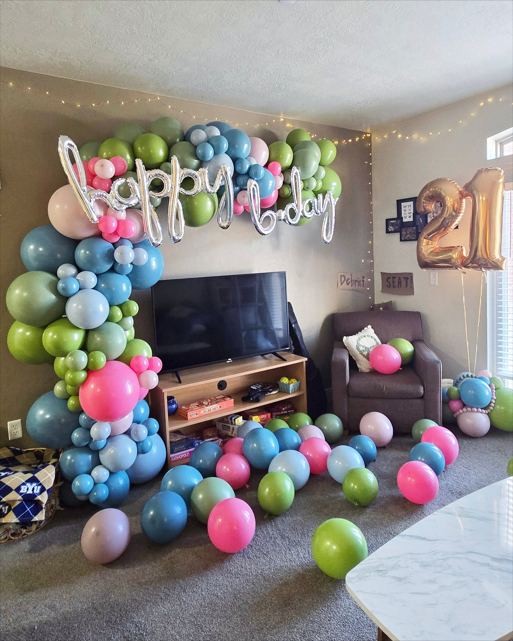 Pink, Green, and Blue balloon garland arches up and over in a living room with the words "happy bday" across the top. Helium number balloons say 21 on the side.