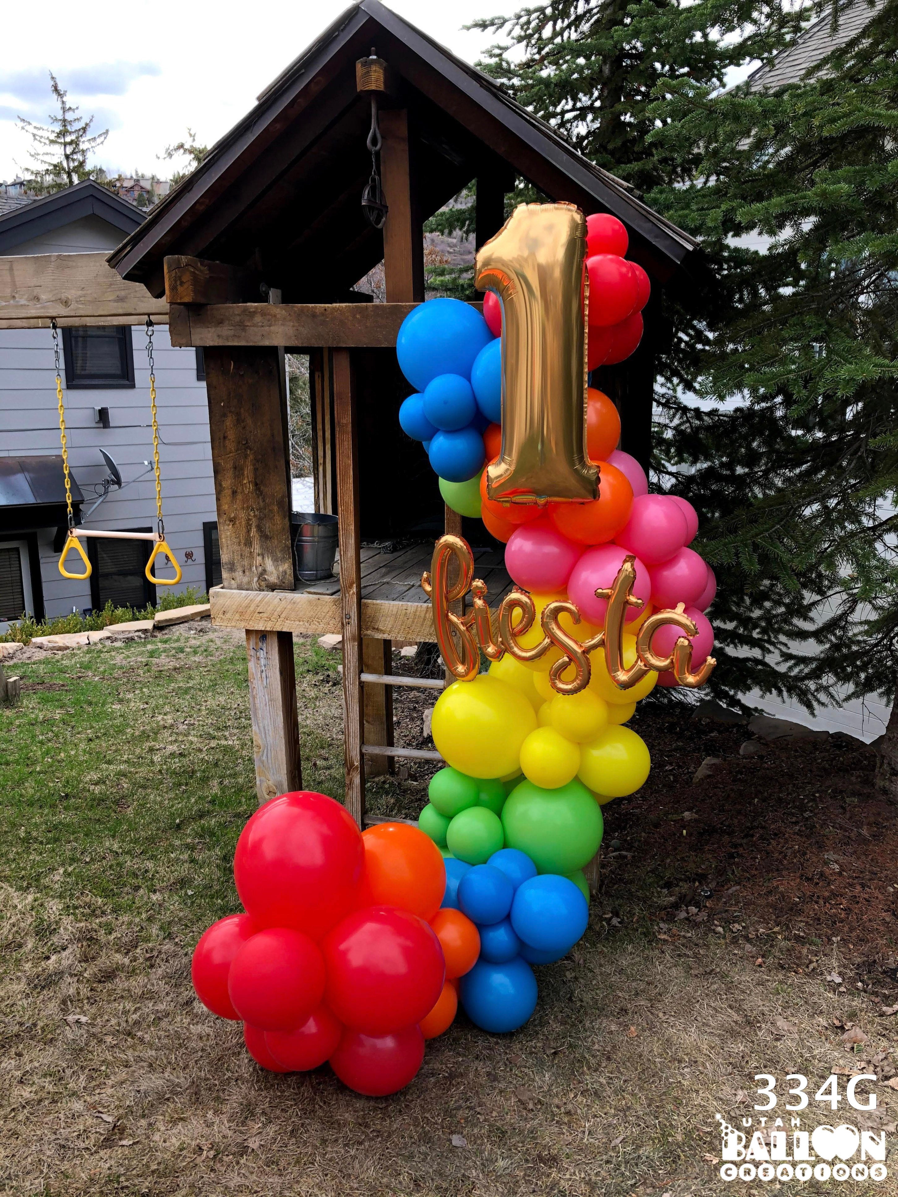 Colorful balloon arch with number '1' and word 'besta' in front of a wooden playhouse.