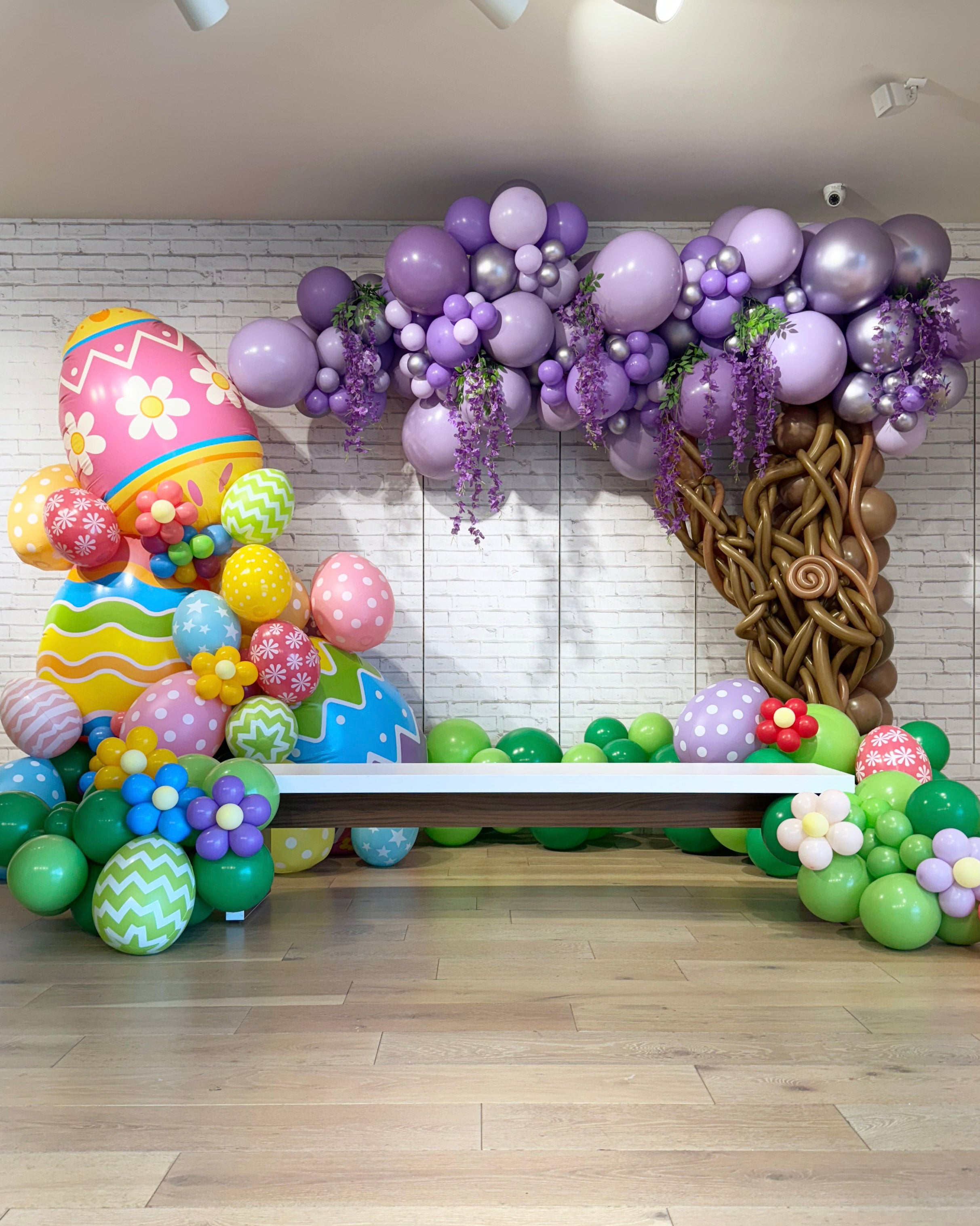 Colorful balloon arch spring tree and easter egg decorations in a room with wooden floor and white wall, setting the stage for pictures with the Easter bunny