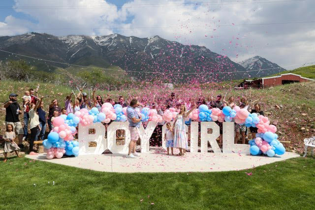 Pink and blue balloons drape across light up letters that say "Boy Girl". People celebrate as pink confetti flies in the air for a gender reveal celebration.