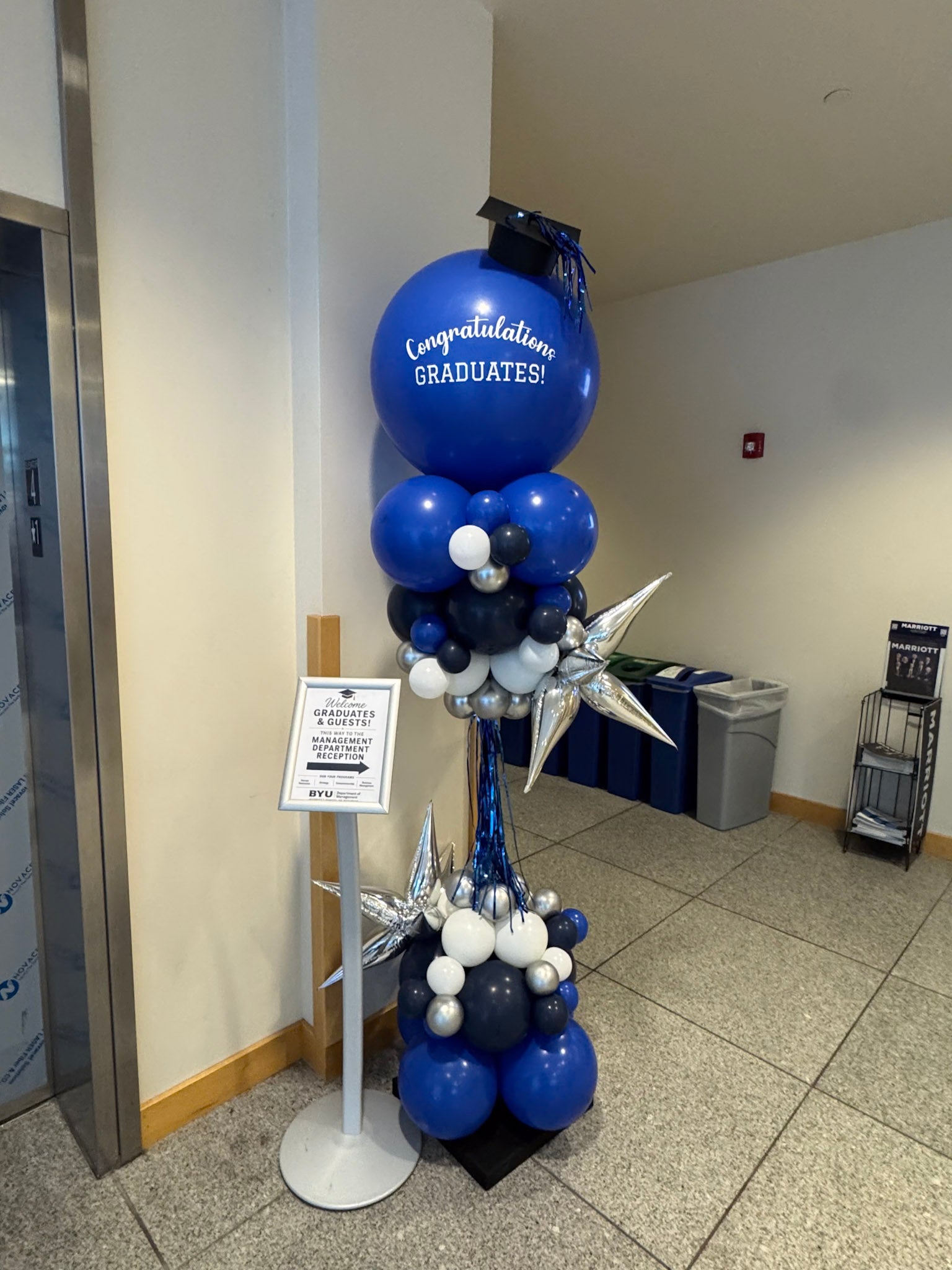 Blue balloon column stands near elevator at BYU marking the entrance for a graduation celebration. Column features "Congratulations Graduates" with a graduation cap and colors royal blue, navy blue, white, silver.