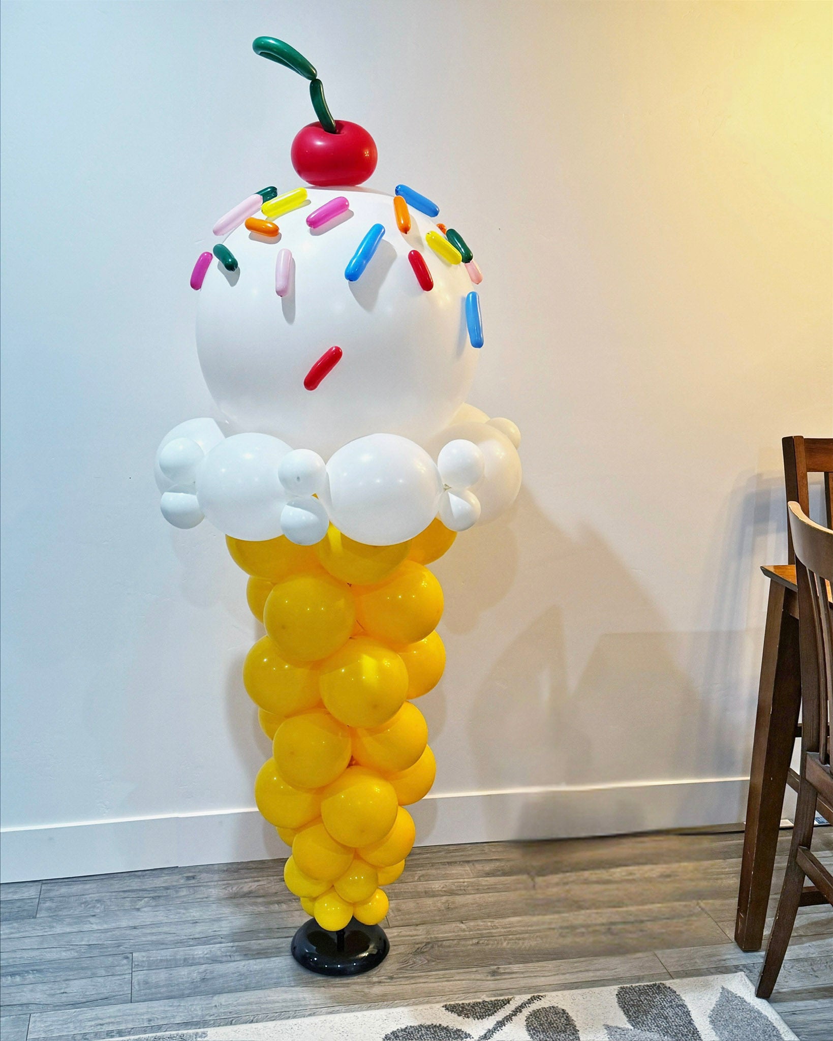 Balloon sculpture of a ice cream cone with colorful sprinkles and a cherry on top against a white wall.