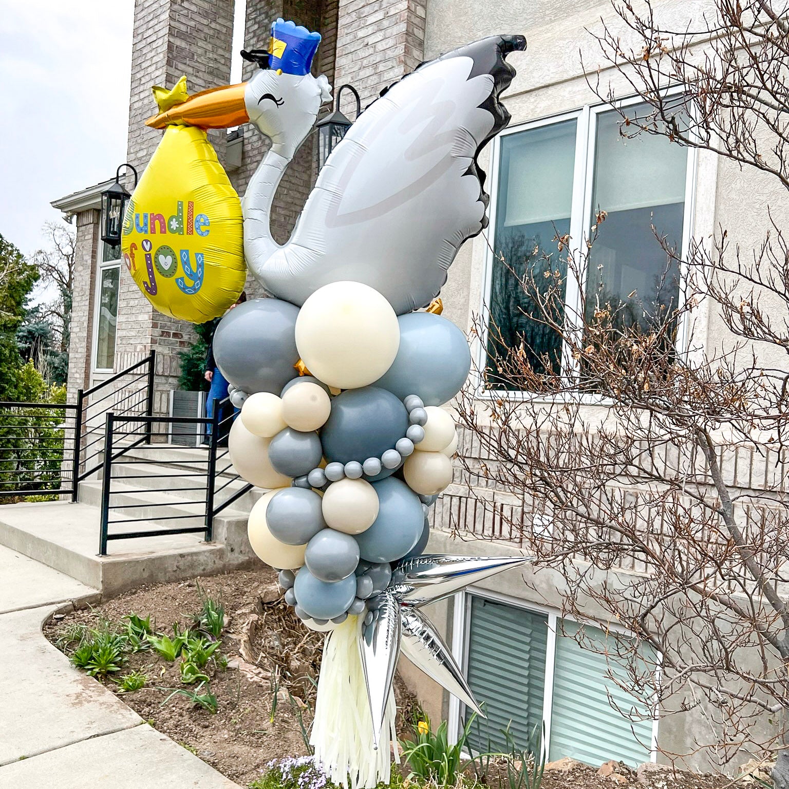 A Stork balloon party pole marks the entrance to the baby shower with light blue and cream balloons outdoors.