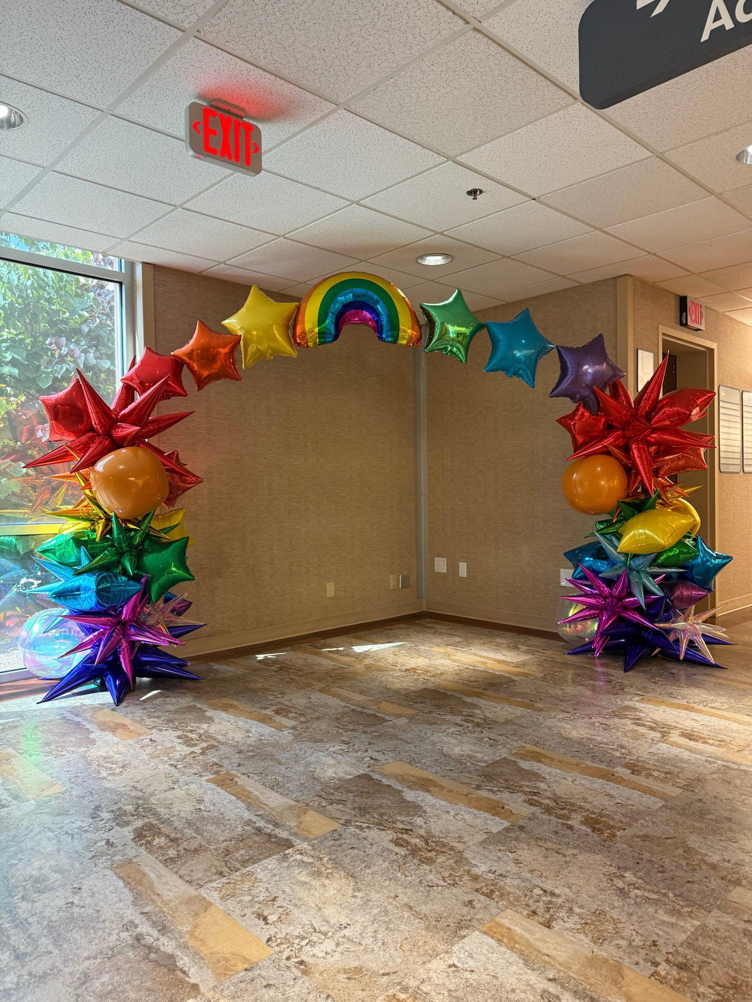 Colorful balloon arch in an indoor setting with a ceiling sign and exit door.