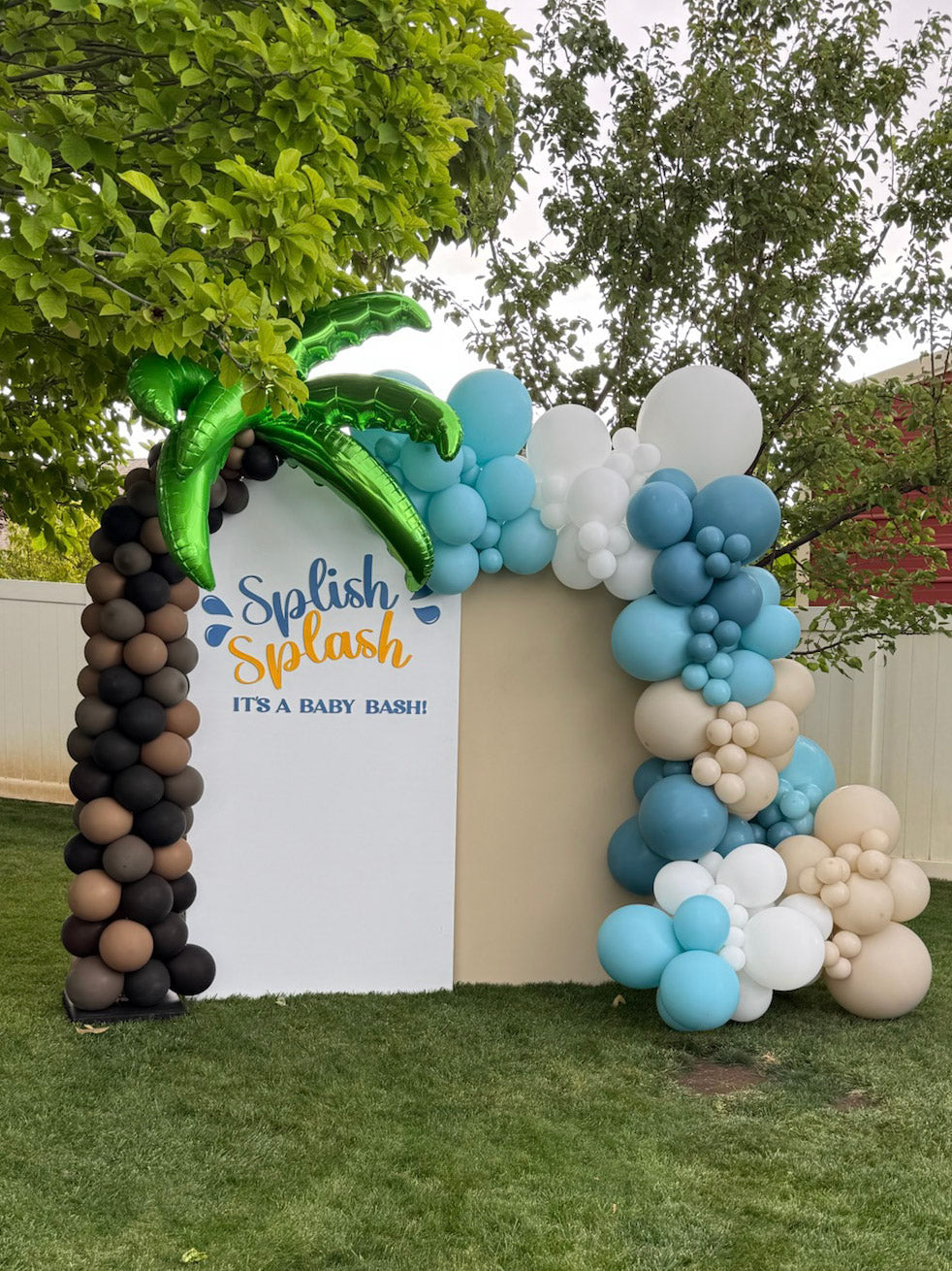 Outdoor backdrop panel for baby shower shows the phrase "Splish Splash it's a baby bash" while balloons create a palm tree sculpture on the left and the right side is framed with a blue and white sand balloon garland.