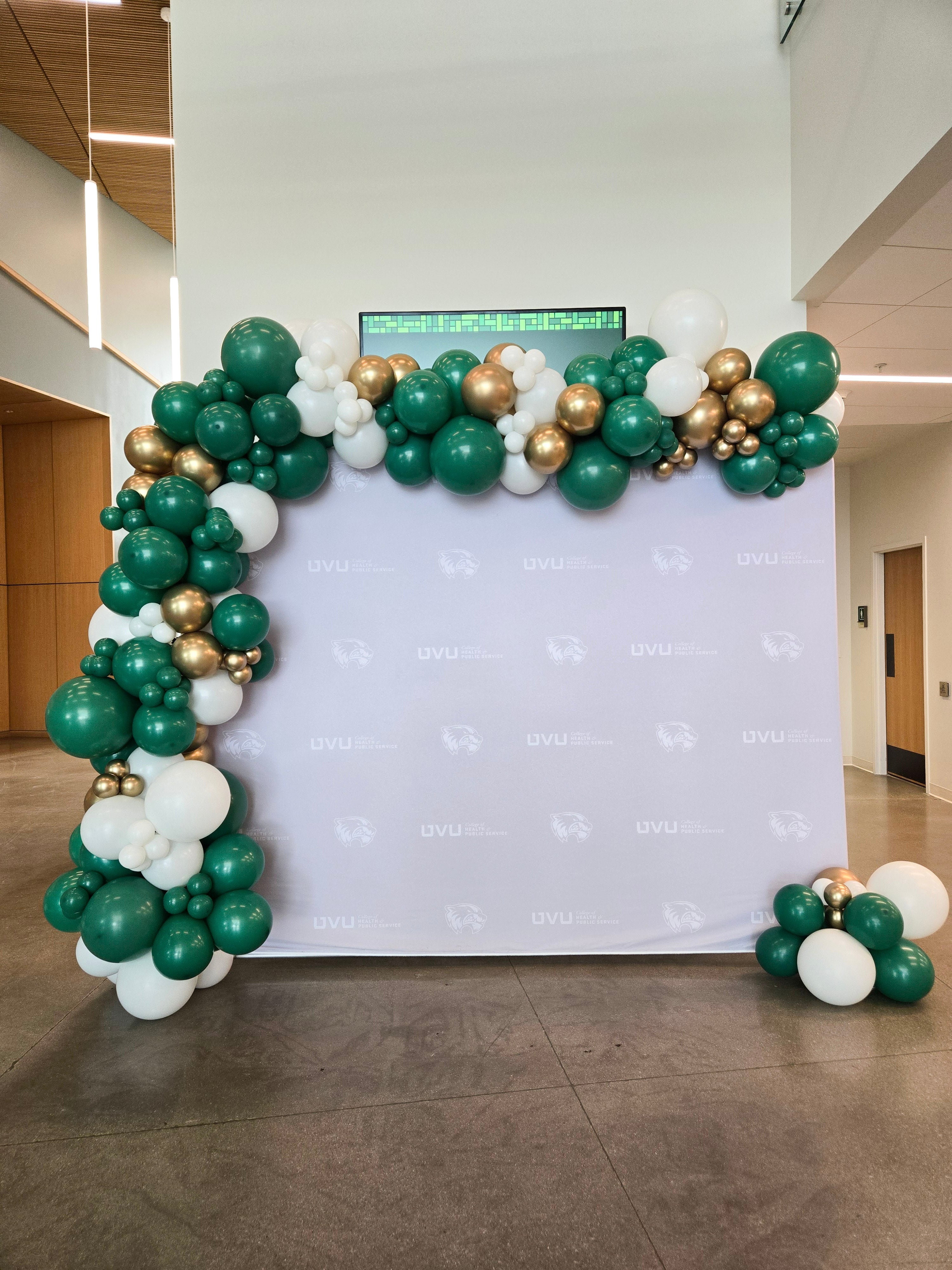 Balloon arch with green, gold, and white balloons in front of a branded backdrop.